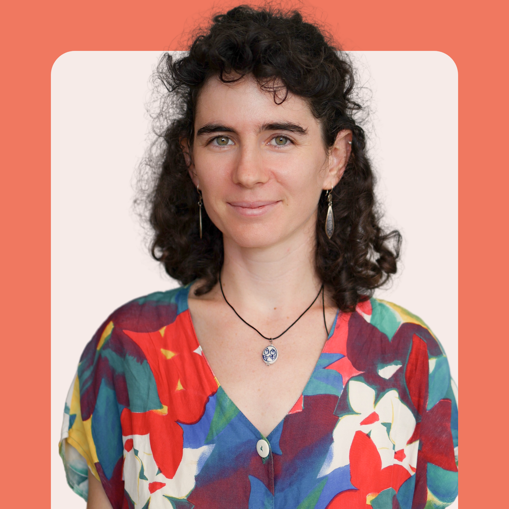 Woman with curly dark hair wearing a colorful floral shirt, drop earrings, and a pendant necklace smiles softly. The background is light pink with a rounded coral border.
