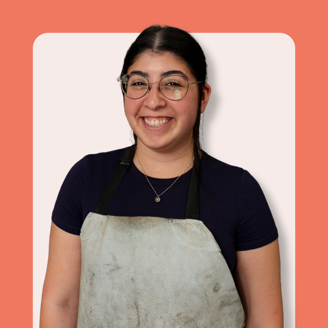 A smiling young woman with glasses and dark hair, wearing a navy shirt and a gray apron, stands in front of a peach and cream background.