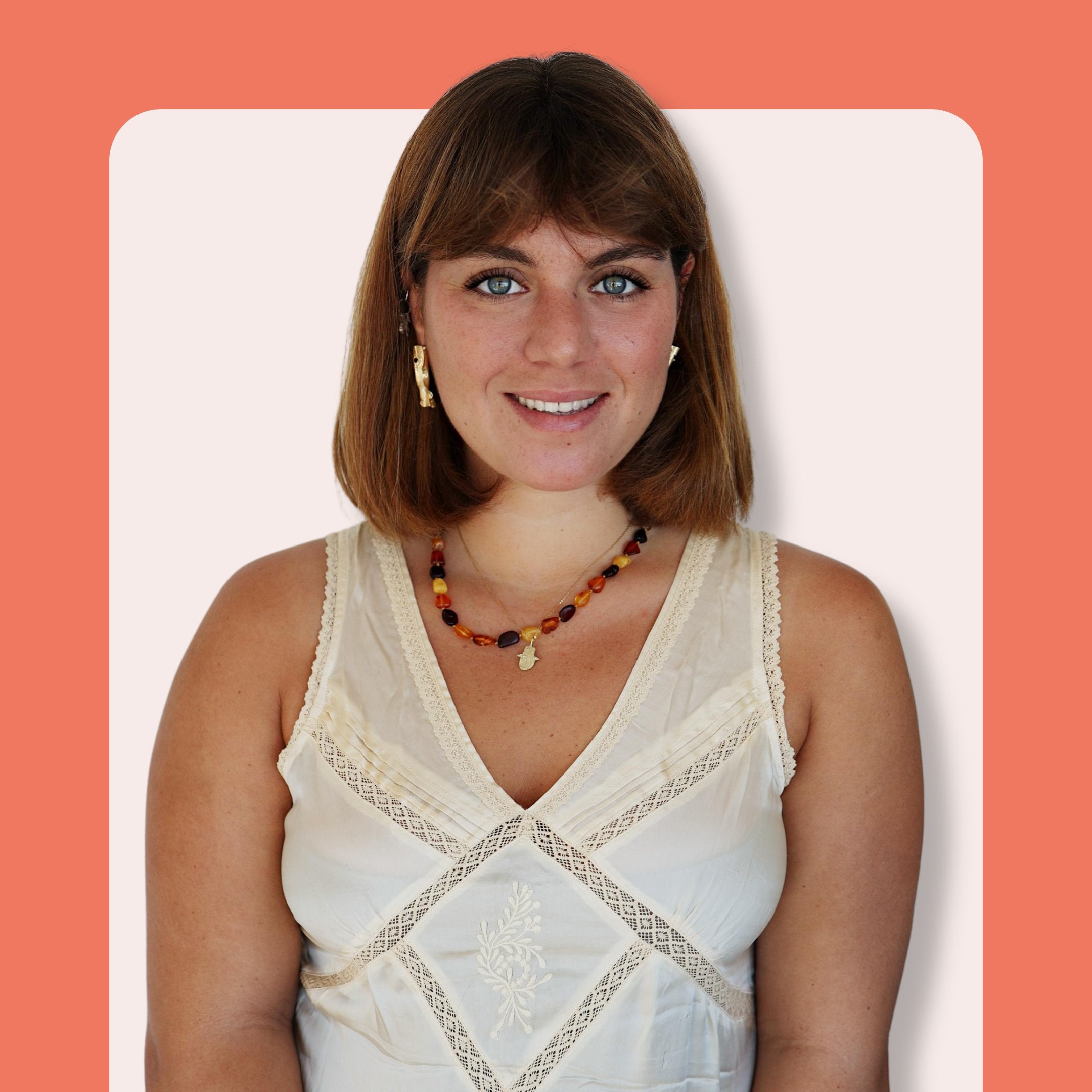 A woman with straight light brown hair, wearing a sleeveless cream lace top, a beaded necklace, and gold earrings, smiles at the camera against a cream and orange background.