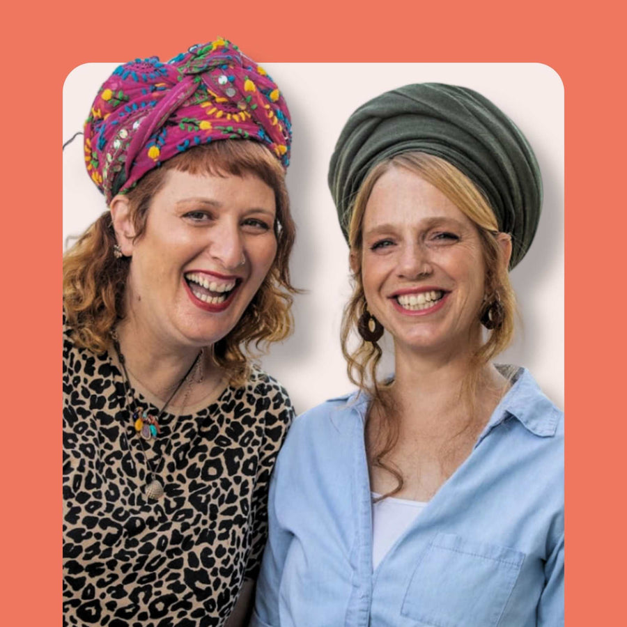 Two smiling women with headwraps, one in a colorful patterned turban and leopard print top, the other in a green headwrap and light blue shirt, standing in front of a peach background.