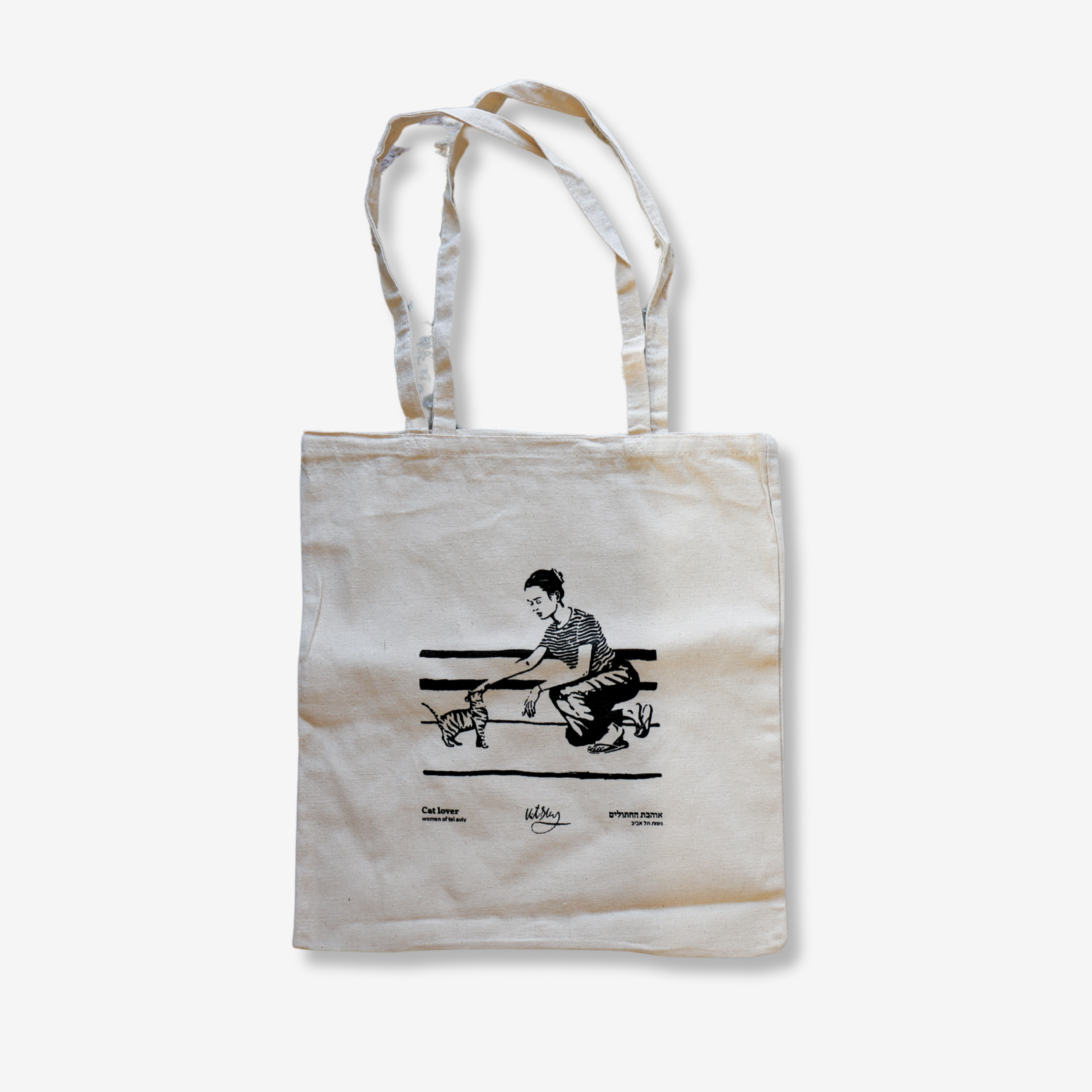 The Cat Lover Tel Aviv Tote Bag by ESEK features a black line illustration of a person petting a cat on a bench in Tel Aviv, with another cat nearby. This cream-colored canvas tote from Vitsky Studio has two handles and text printed below the design.