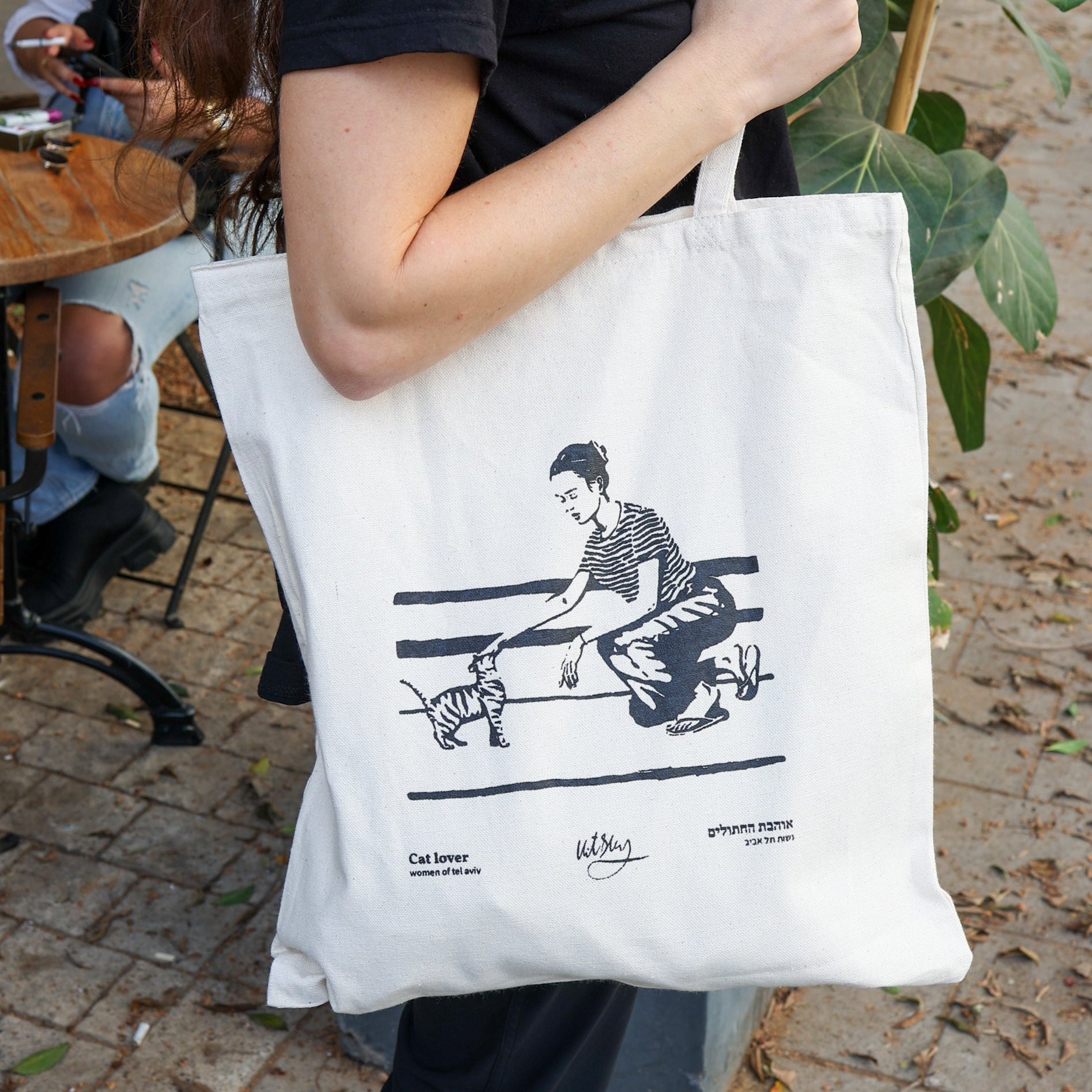 A person holds the Cat Lover Tel Aviv Tote Bag by ESEKs Vitsky line, featuring a black line drawing of someone petting a cat and text in English and Hebrew, outdoors near Tel Aviv by a wooden table and lush plants.