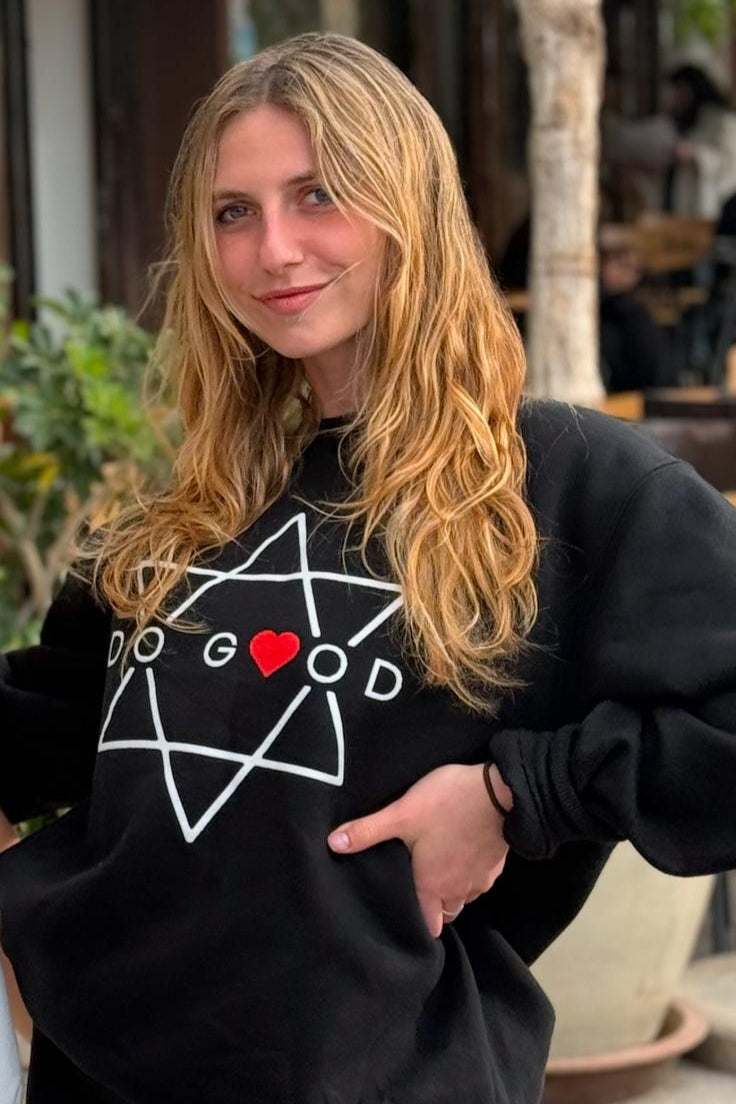 A smiling woman with long blonde hair wears the ESEK Sword of Iron Volunteer Corp. Do Good Sweater, featuring a white star, DO GOOD text, and a red heart, standing outdoors against greenery with a blurred background.