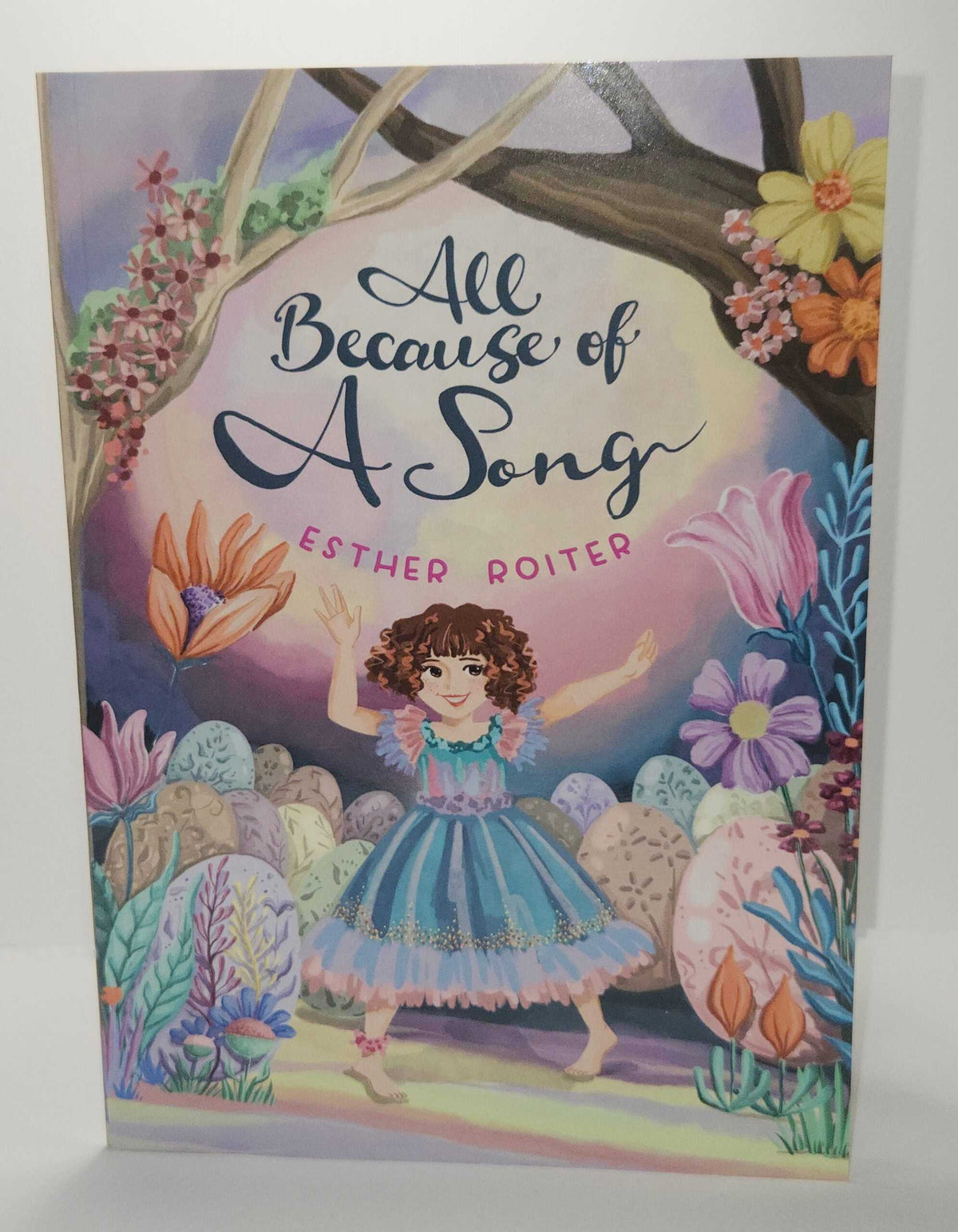 ADI All Because of a Song children’s book with a colorful illustrated cover, featuring a girl in a blue dress among flowers and trees.