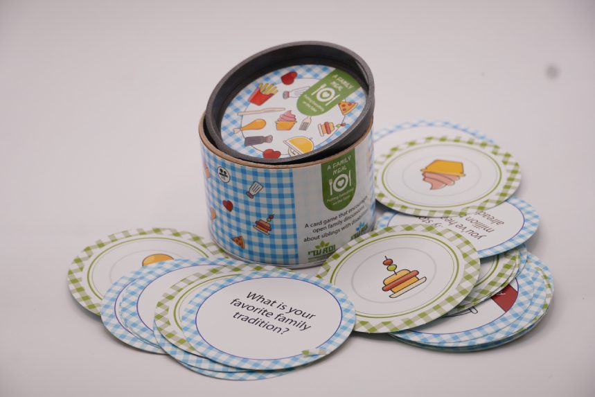 ADI A Family Meal Card Game in a blue gingham cylindrical tin with colorful illustrated round cards featuring food and family prompts.