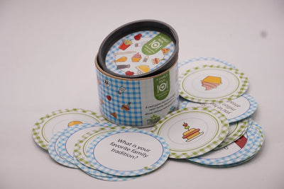 ADI A Family Meal Card Game in a blue gingham cylindrical tin with colorful illustrated round cards featuring food and family prompts.