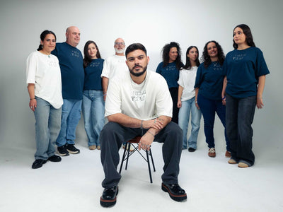Omer Wenkert Now Is Good T-shirt in white cotton, with Hebrew lettering on the chest, modeled by a group of people.